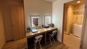 Quarto compacto com área de toucador, espelho com iluminação, duas cadeiras, mesa pequena com planta e guarda-roupa.