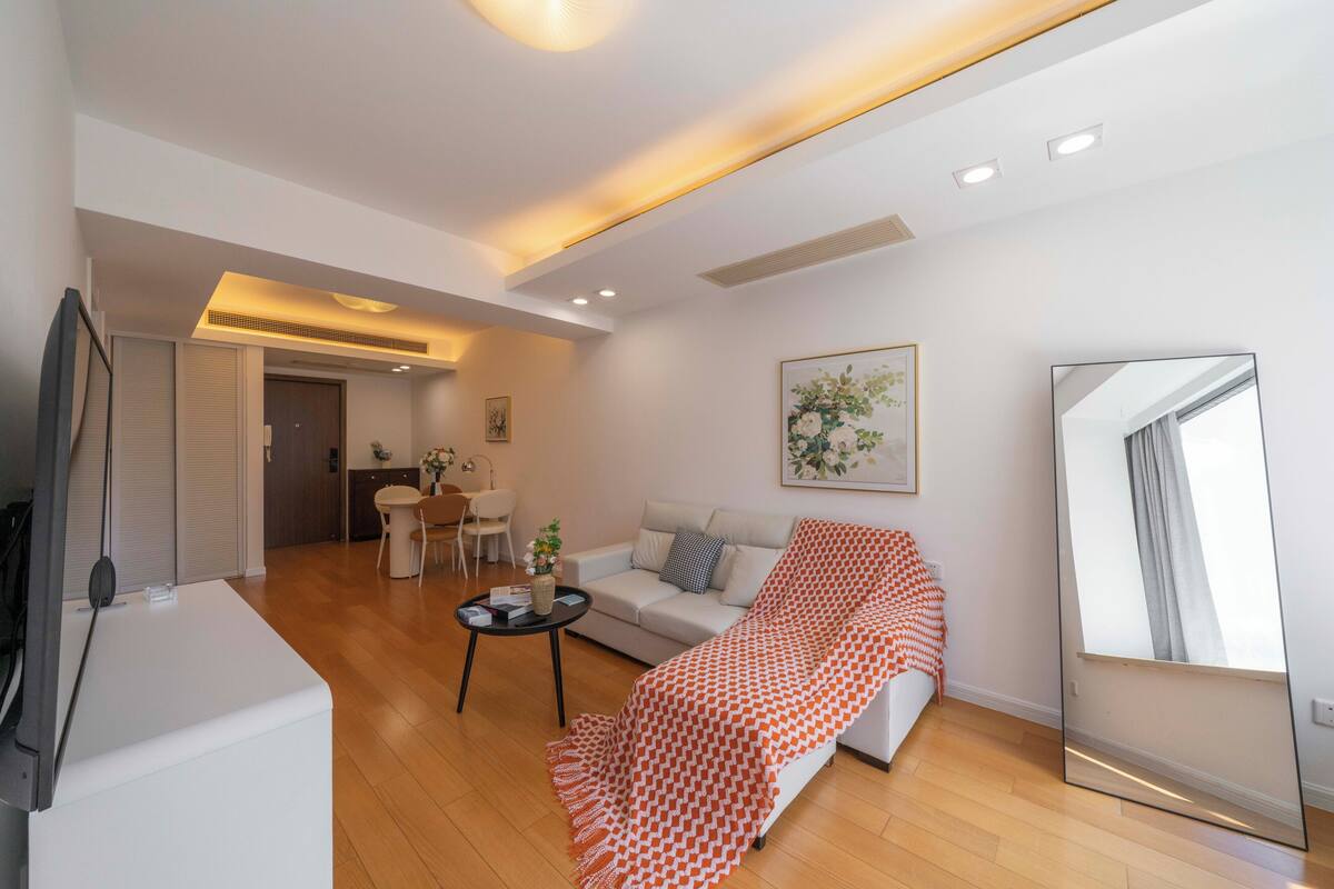 A modern living room with a sofa, a coffee table, and a TV. There is a dining area in the background with a table and chairs.