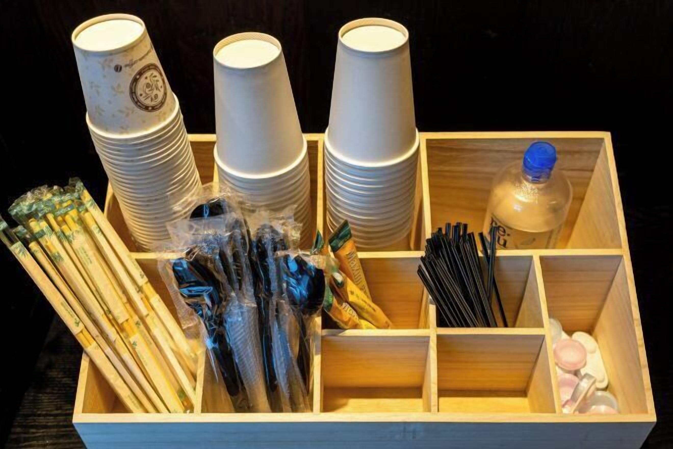 A wooden box containing stacks of white paper cups, plastic utensils, and a bottle of water.