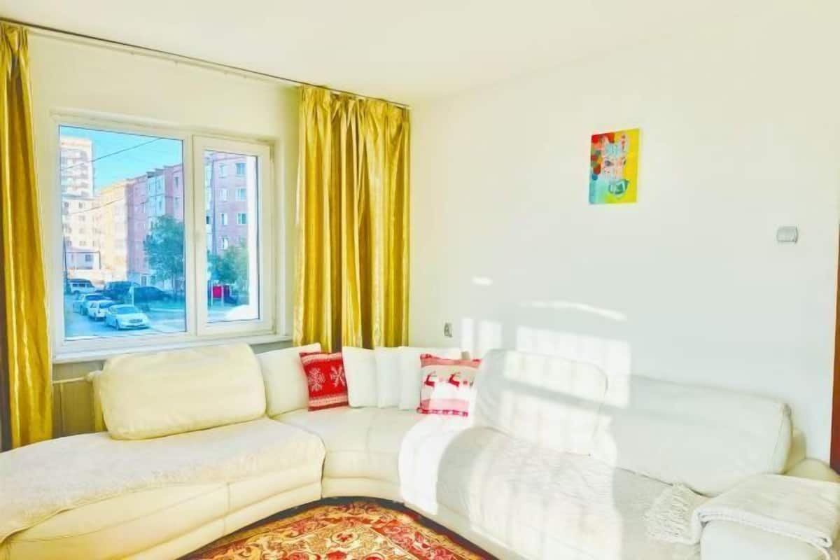 A living room with a white sofa, a patterned rug, and a window with yellow curtains.