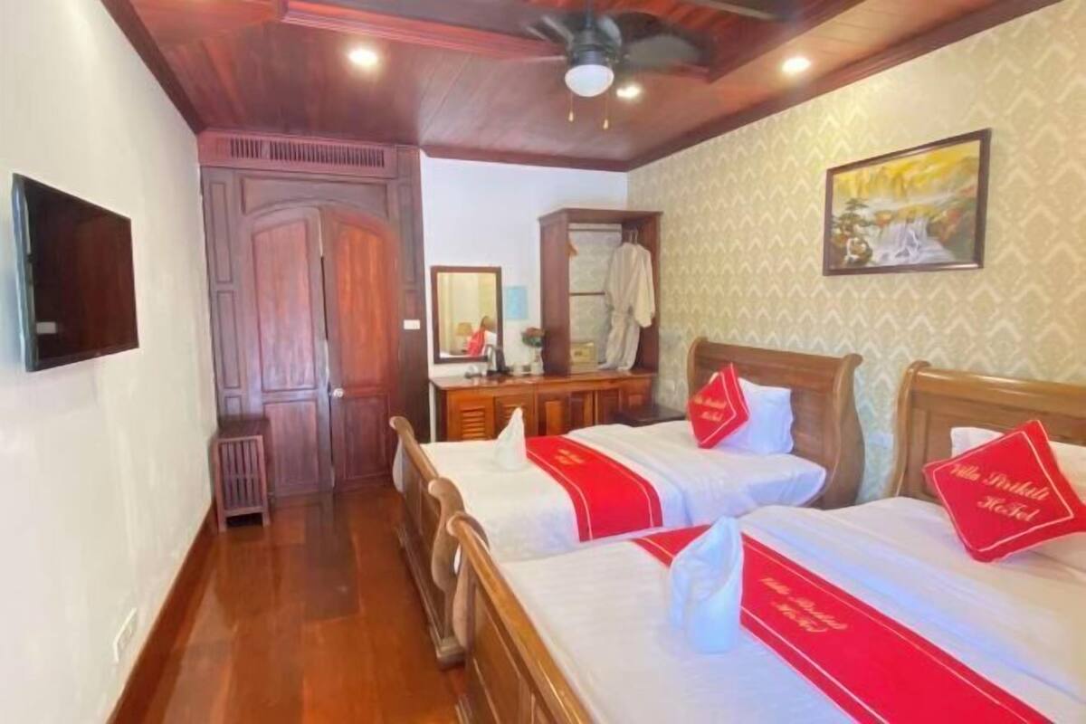 Une chambre d’hôtel avec deux lits, une armoire en bois, un miroir et un ventilateur de plafond.