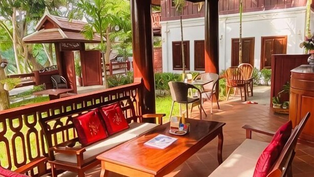 A wooden outdoor seating area with a table and chairs, a wooden bench with cushions, and a view of a building with a red roof and a white wall.