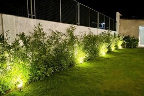Un espace jardin bien éclairé, avec un mur blanc et une clôture en métal, où l’herbe et les plantes sont soigneusement taillées.