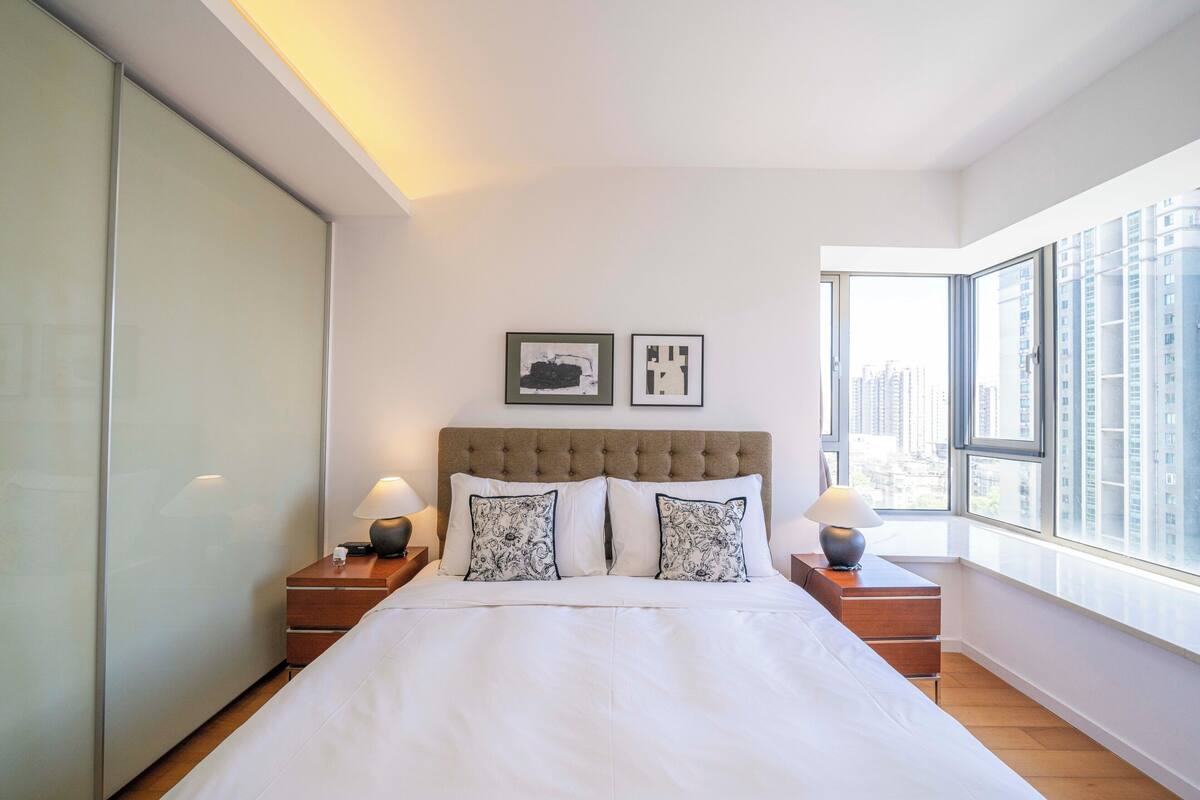Une chambre moderne avec un grand lit, deux tables de chevet, une fenêtre donnant sur la ville et deux tableaux encadrés accrochés au mur.