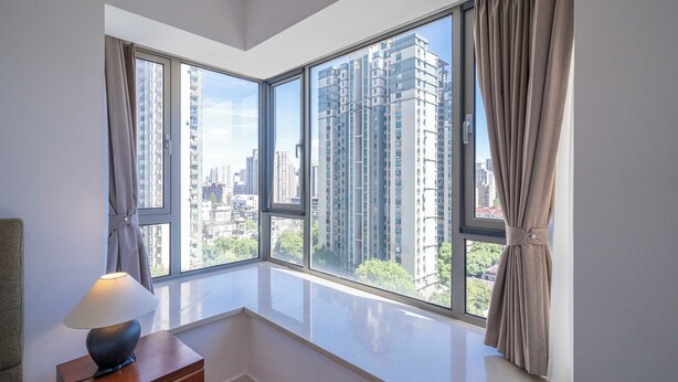 A modern room with a large window, a bedside table with a lamp, and a view of high-rise buildings. - Tyms Elegant 2BR 1BA Apt Walk to Malls (Shanghai)