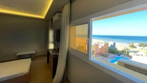 A hotel room with a bed, a desk, a TV, and a window with a view of the beach and pool. - Al Dora All Inclusive Hotel (Hurghada)