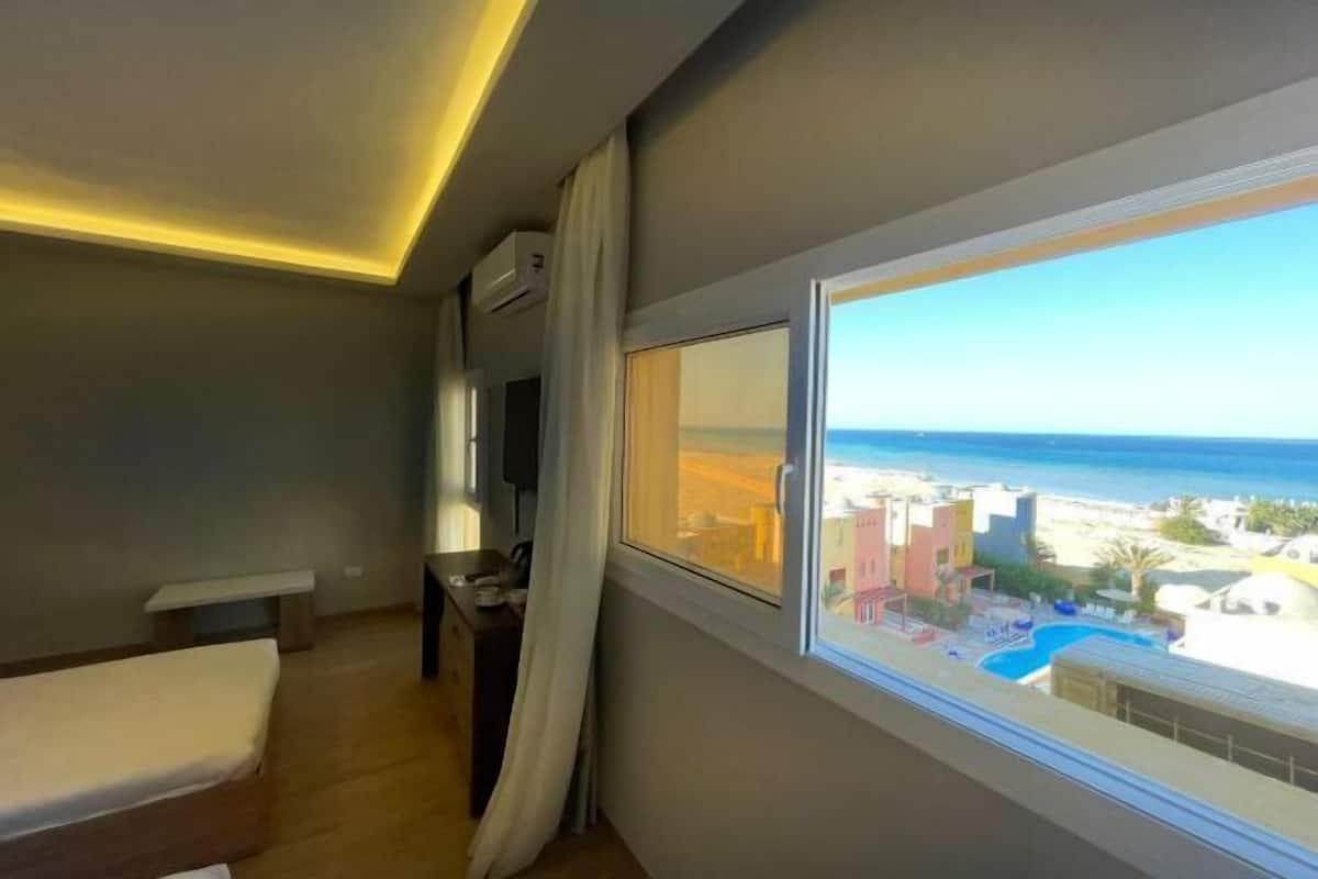 Ein Hotelzimmer mit Bett, Schreibtisch, Fernseher und Fenster mit Blick auf Strand und Pool.