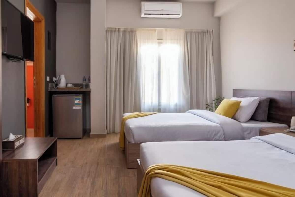 Ein Hotelzimmer mit zwei Betten, einem kleinen Schreibtisch, einem Kühlschrank und einem Fenster mit Vorhängen.