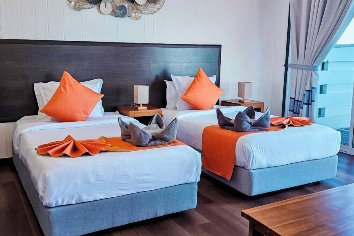 A hotel room with two beds, each with a dark headboard, white bedding, and orange pillows. There are bedside tables with lamps and a view of the city through large windows.
