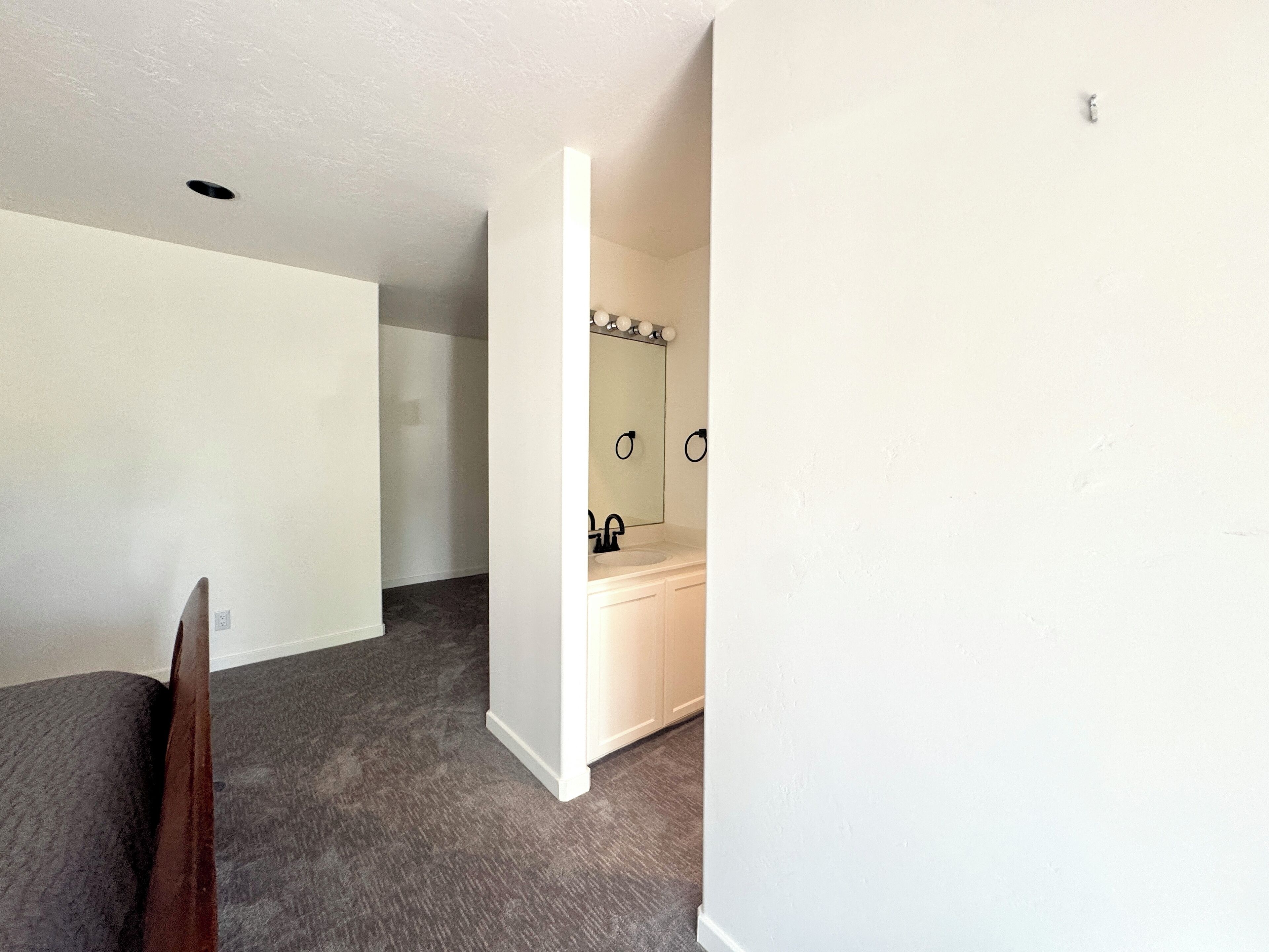 Ein Zimmer mit grauem Teppich, weißen Wänden, einem Holzstuhl und einem Badezimmer mit Spiegel, Waschbecken und Handtuchhalter.