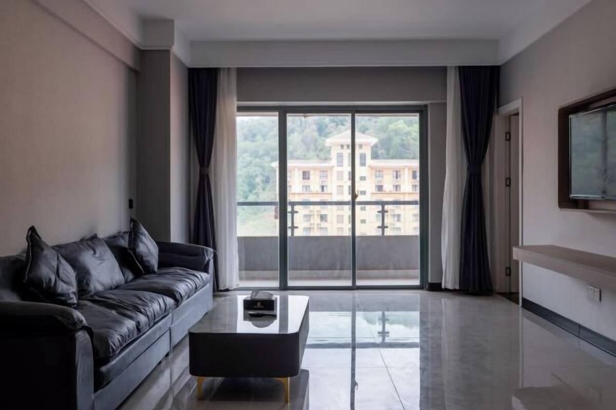 A modern living room with a black leather sofa, a glass coffee table, and a large sliding glass door leading to a balcony with a view of a building.