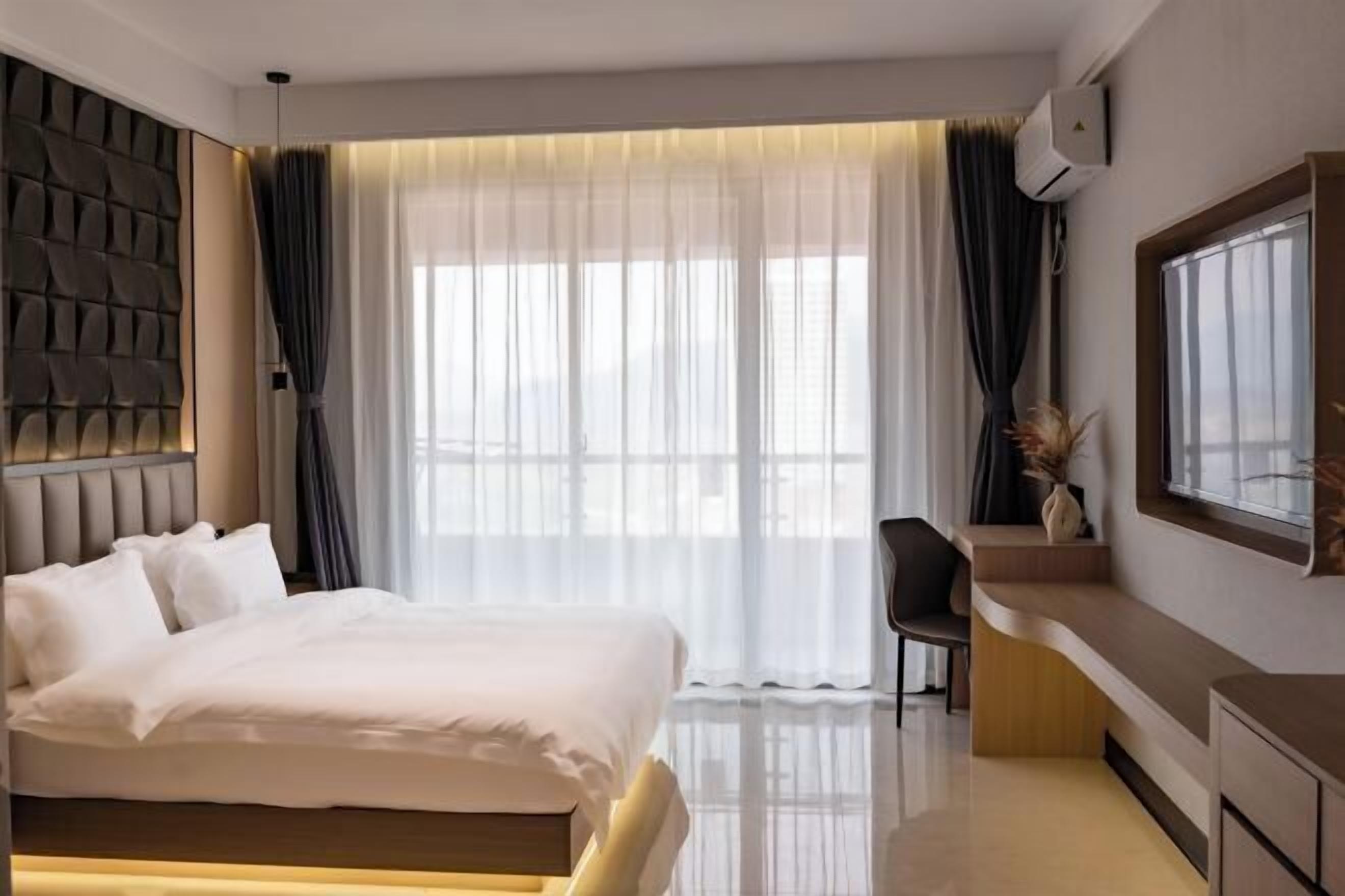 A modern hotel room with a large bed, a desk, and a window with sheer curtains.