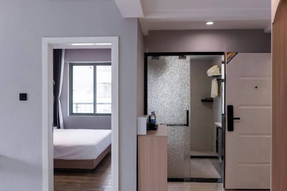 A modern hotel room with a bed, a bathroom with a shower, and a door leading to another room.