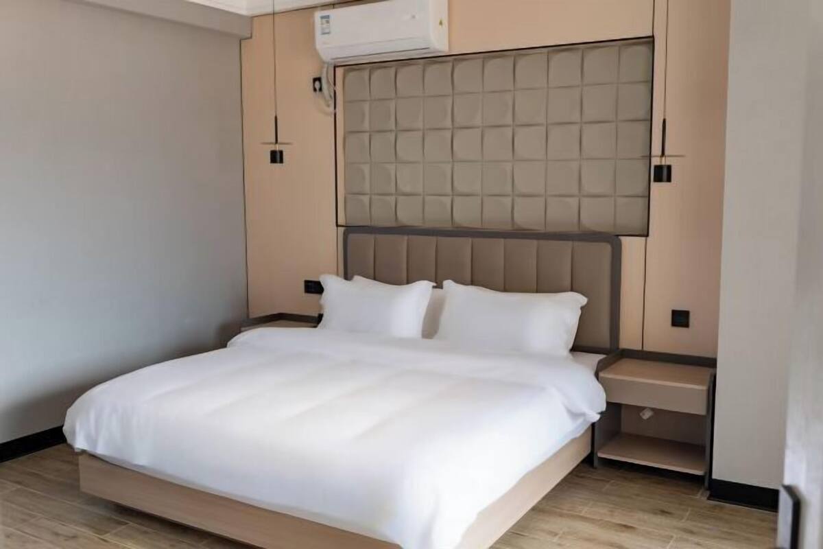 A modern hotel room with a large bed, a padded headboard, a bedside table, and an air conditioning unit.