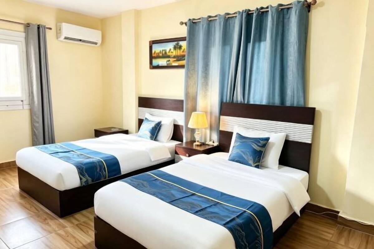 A hotel room with two beds, a nightstand, a lamp, an air conditioning unit, and a painting on the wall.