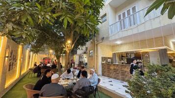 An outdoor dining area with tables and chairs, surrounded by greenery and a building with a balcony.