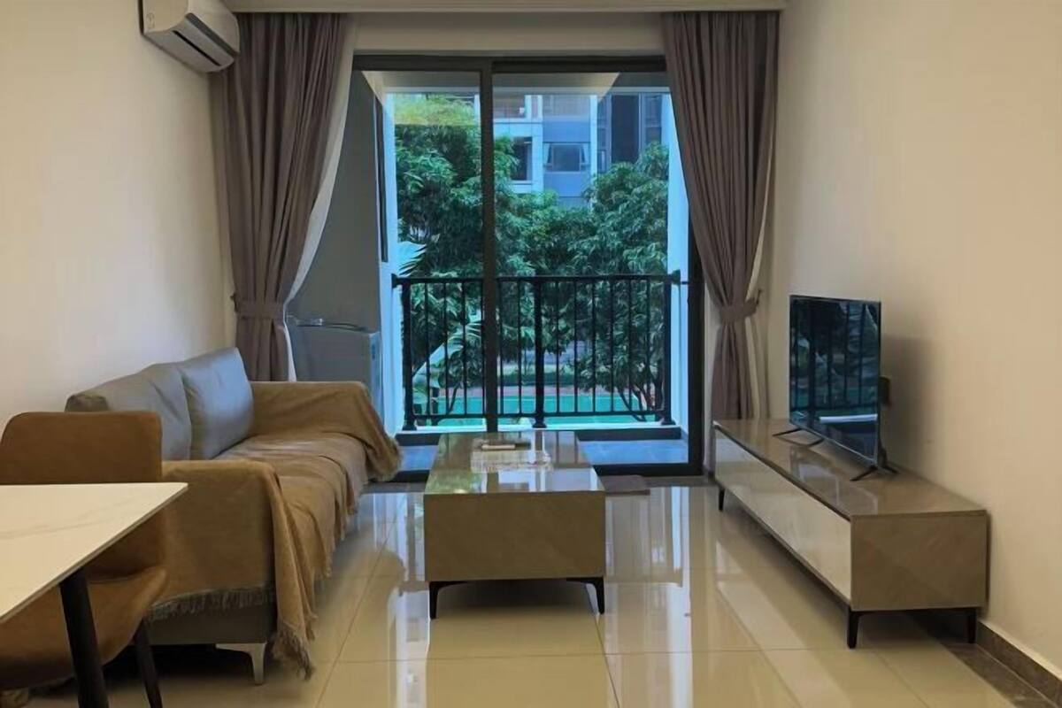 A modern living room with a sofa, a coffee table, a TV, and a balcony with a view of greenery.