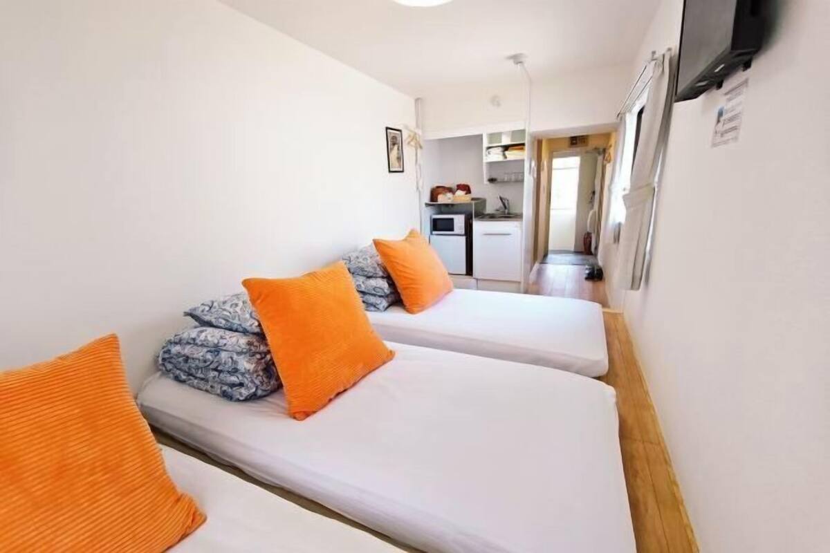 A compact room with two beds, each with an orange pillow, a small kitchenette, and a television mounted on the wall.