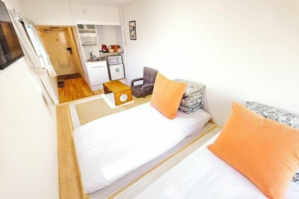 A compact hotel room with a bed, a small desk, a chair, and a kitchenette.