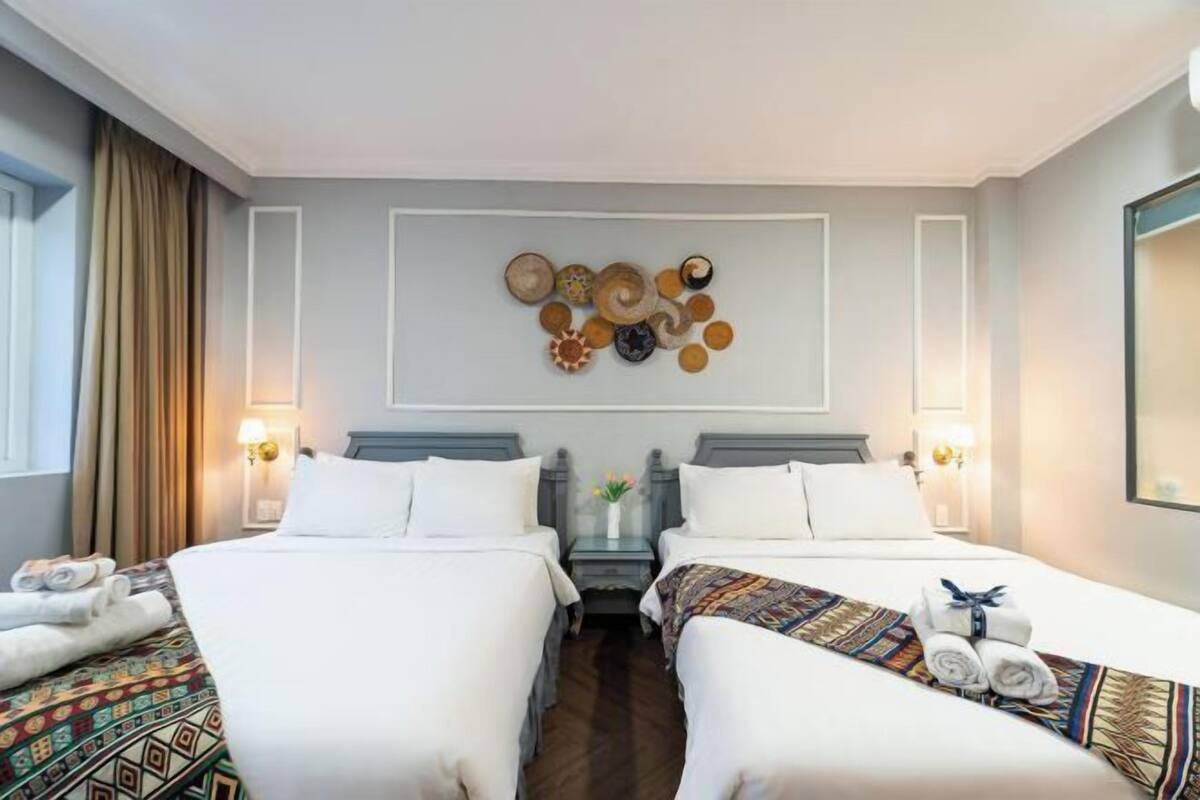 A hotel room with two beds, a decorative wall art piece, a nightstand with a lamp, and a window with curtains.