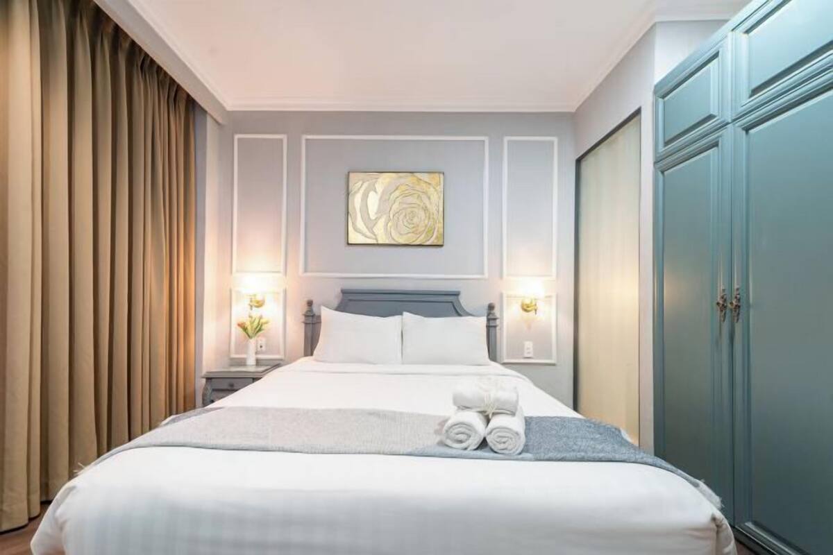 A neatly made bed with white linens, a grey throw blanket, and rolled towels on the bed. There is a wall-mounted artwork above the bed, two bedside lamps, and a wardrobe with a blue door.