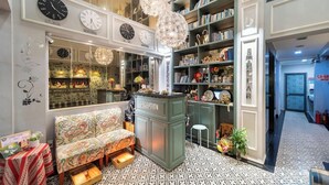 Zona de recepción con un mostrador verde, varios relojes, un sofá estampado y una estantería llena de libros y objetos decorativos.