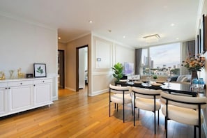 A modern hotel room with a wooden floor, a dining area with a table and chairs, and a view of the cityscape through large windows.