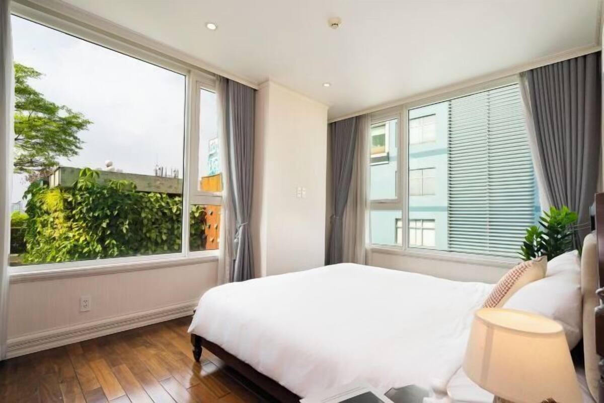 A bedroom with a large window, a bed, a bedside table, and a lamp.