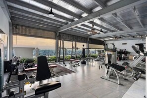 Une salle de sport équipée de divers appareils d’exercice, une grande fenêtre offrant une vue sur la ville et un plafond haut avec des poutres apparentes.