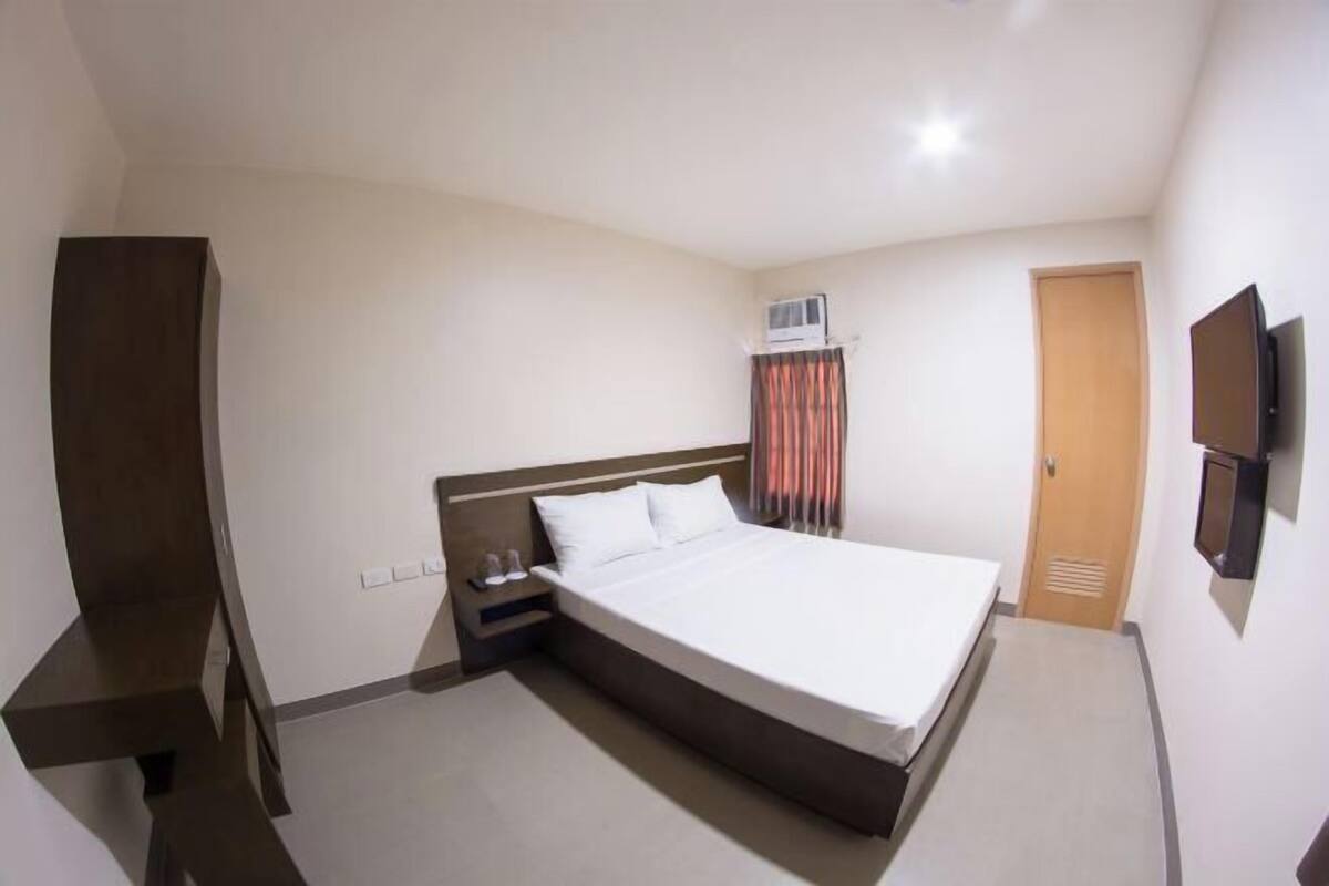 A hotel room with a bed, a TV, a wardrobe, a bedside table, and an air conditioning unit.