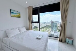 Ein Schlafzimmer mit Bett, einem Fenster mit Vorhängen, einem kleinen Nachttisch und Blick auf die Stadt.