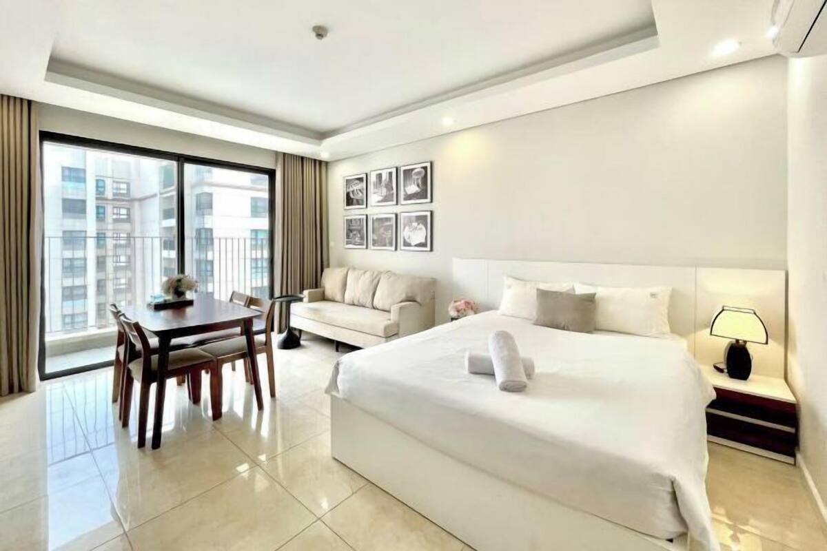 Ein Hotelzimmer mit einem großen Bett, einem Esstisch mit Stühlen, einer Couch, einem Nachttisch mit Lampe und Blick auf die Stadt durch große Fenster.
