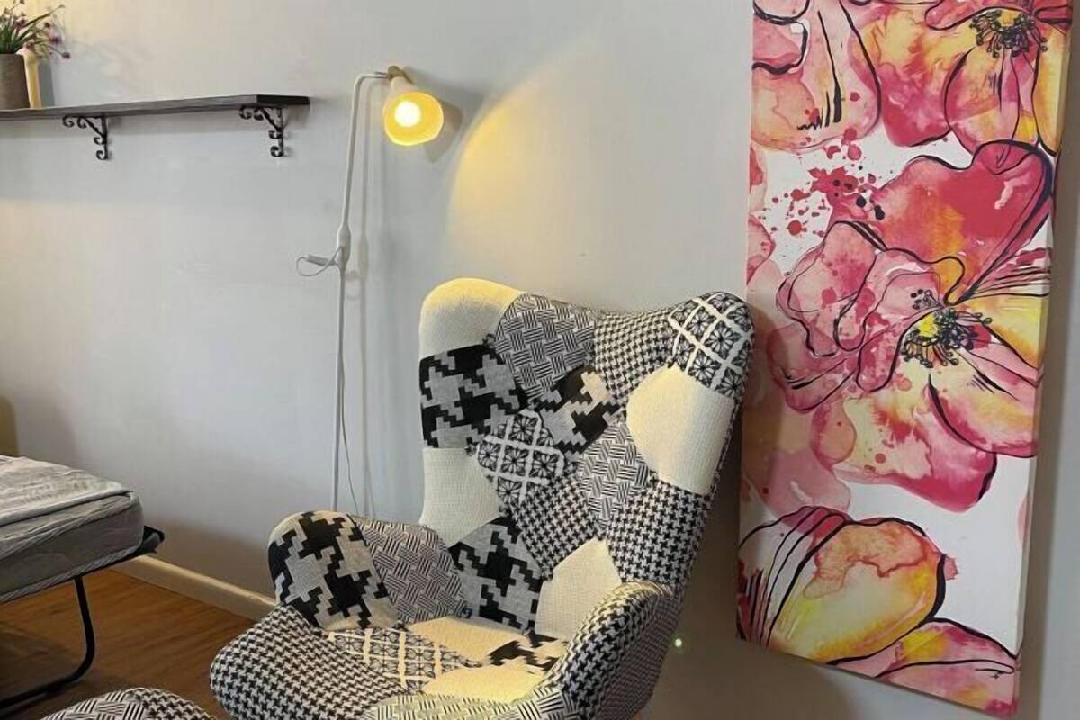 A chair with a patchwork design, a bedside table, a lamp, and a floral painting on the wall.
