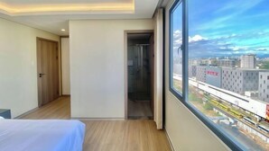 A modern hotel room with a large window, a bed, a door, and a view of the cityscape.