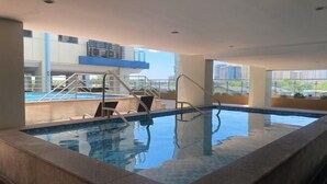 An indoor swimming pool with clear blue water, a diving platform, and a view of the cityscape through large windows.