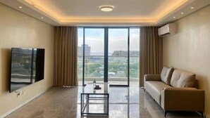 A modern living room with a large flat-screen TV, a sofa, a coffee table, and a view of a cityscape through large windows.