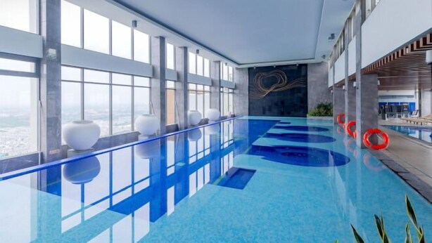 A modern indoor swimming pool with a glass barrier, large windows, and a view of the city. - A Quiet Stay - Service Apartment, Vinhome Ocean Park, Ha Noi (Hanoi)