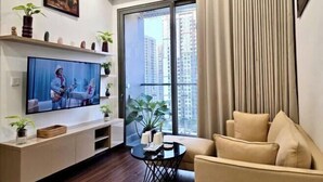 A modern living room with a flat-screen TV, a sofa, a small coffee table, and a balcony with a view of high-rise buildings. - A Quiet Stay - Service Apartment, Vinhome Ocean Park, Ha Noi (Hanoi)