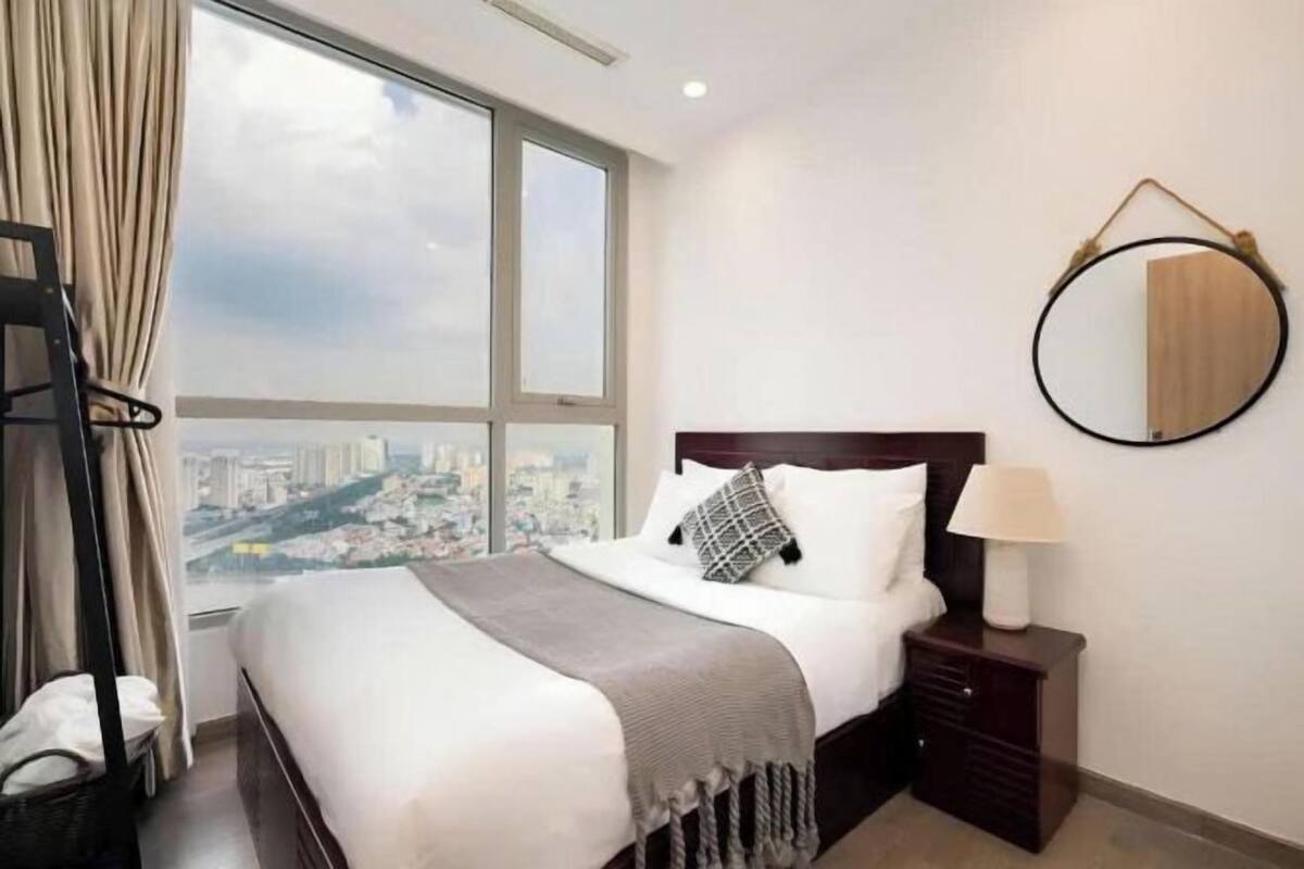 Una habitación de hotel con un ventanal grande, una cama con edredón blanco, cabecera de madera, mesita de noche con lámpara y un espejo redondo en la pared.