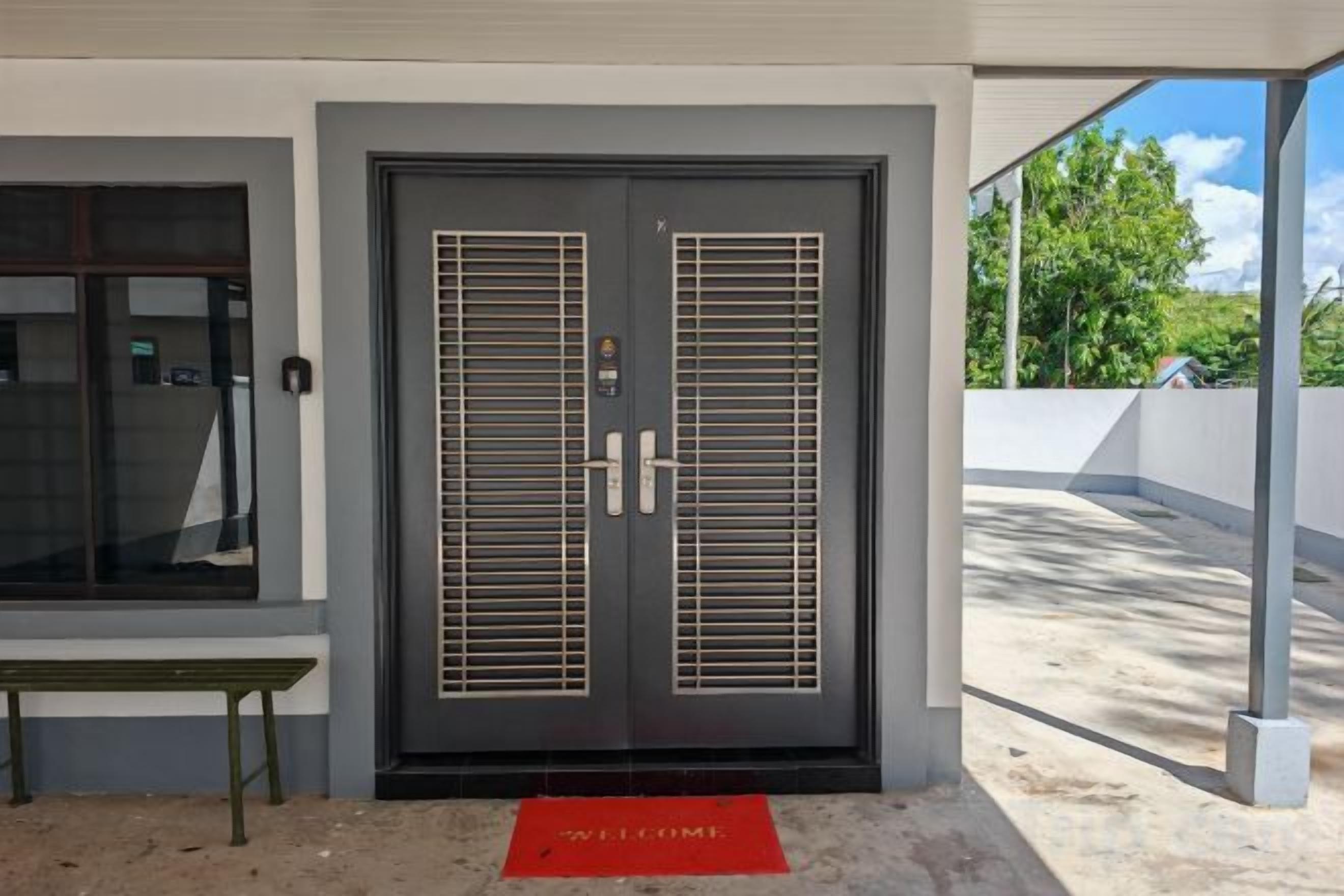 Une porte à deux battants équipée d’un clavier de sécurité, un banc et un tapis rouge sur lequel est écrit le mot « BIENVENUE ».