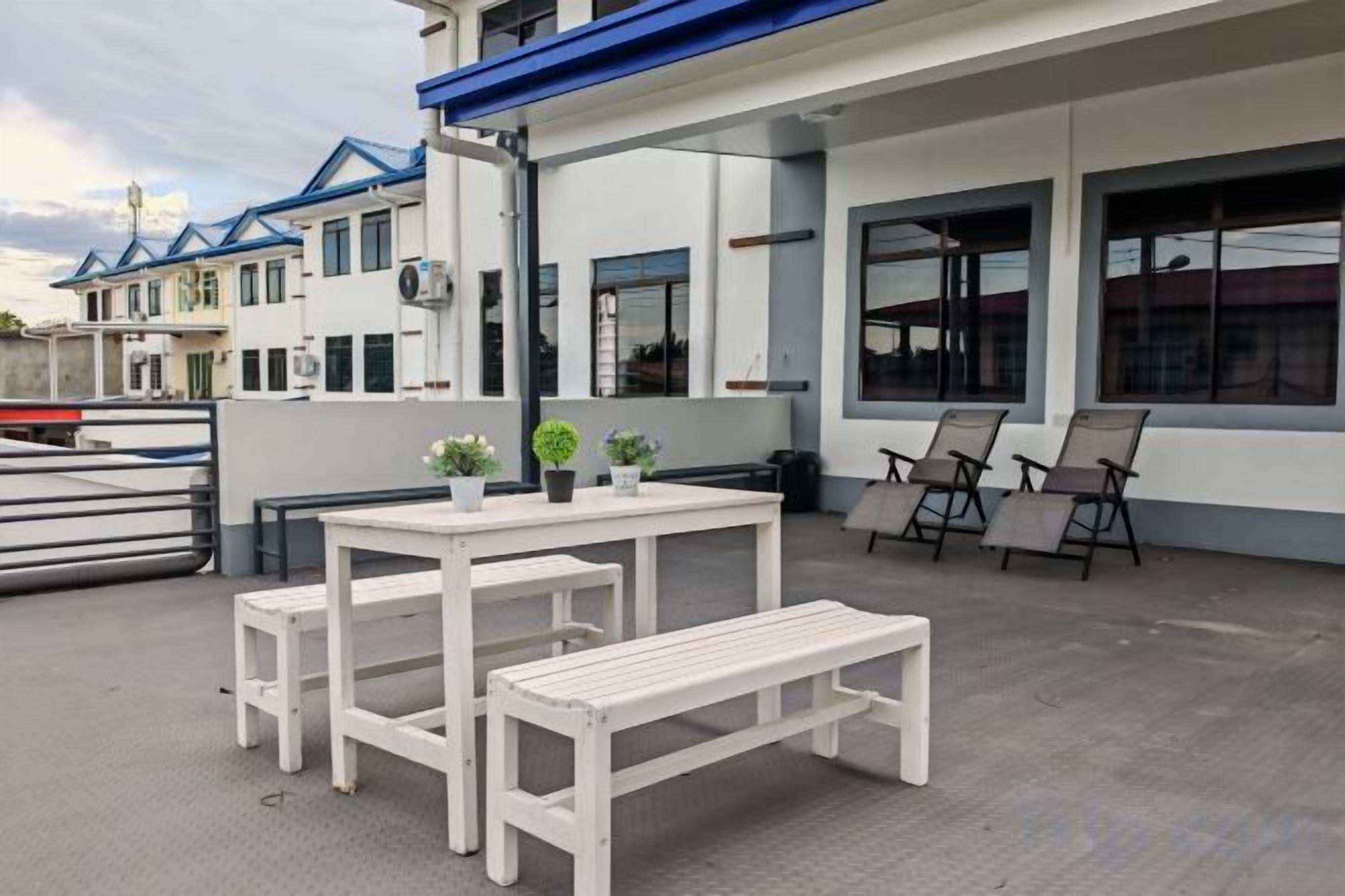 An outdoor seating area with a table and benches, a trash bin, and a building with large windows and a blue roof.