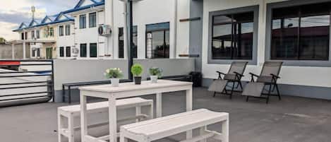 An outdoor seating area with a table and benches, a trash bin, and a building with large windows and a blue roof.