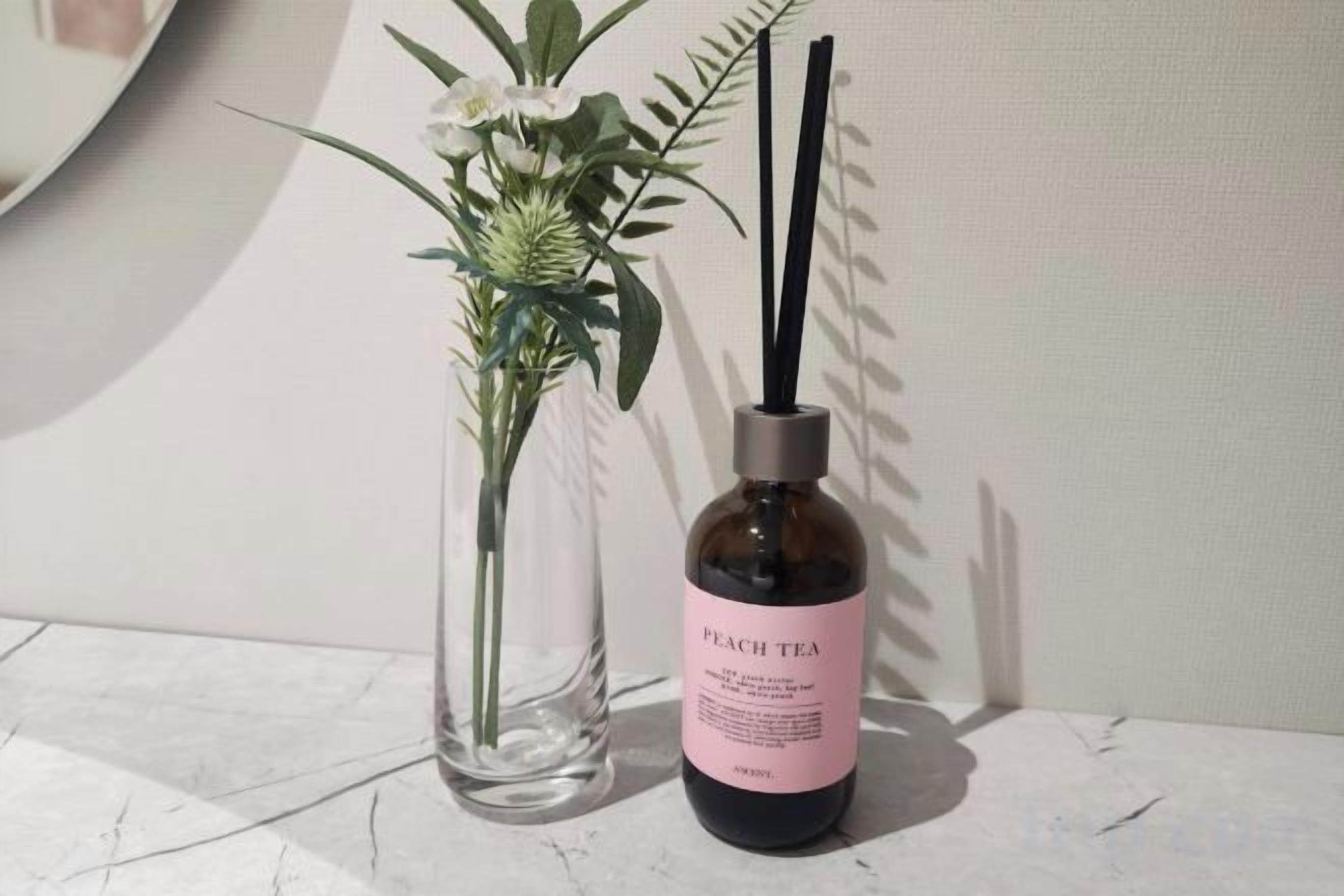 Un florero de vidrio transparente con un arreglo floral y una botella etiquetada 'PEACH TEA' con tapón negro y un difusor de caña.