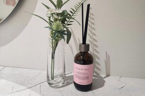 En gennemsigtig glasvase med blomsterarrangement og en flaske mærket 'PEACH TEA' med sort låg og en diffuserpind.