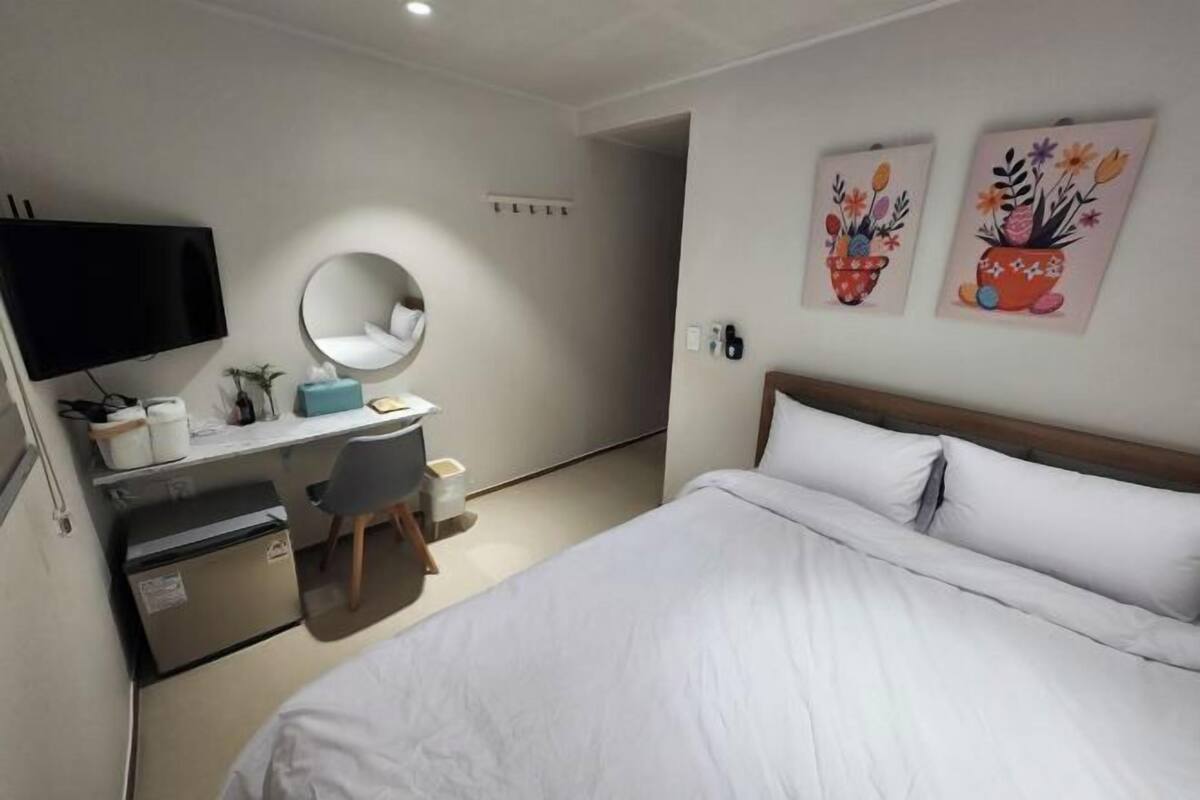 A hotel room with a bed, a desk with a chair, a television, a refrigerator, and two wall-mounted paintings.