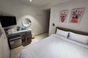 Ett hotellrum med en säng, ett skrivbord med en stol, en tv, ett kylskåp och två tavlor som hänger på väggen.