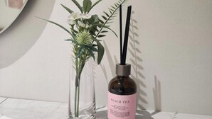 Un vase en verre transparent contenant des fleurs blanches, et une bouteille noire avec une étiquette rose sur laquelle est inscrit « THÉ À LA PÊCHE », placés côte à côte.