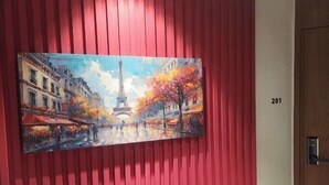 Une peinture représentant la Tour Eiffel dans une scène de rue parisienne, accrochée à un mur rouge avec une porte marquée du numéro « 201 » sur le côté droit.