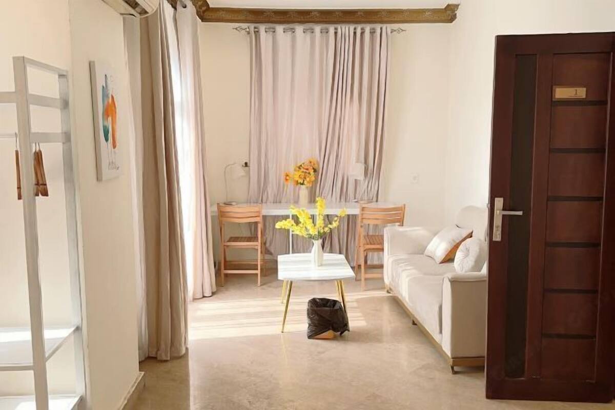 A living room with a white sofa, a coffee table with a vase of flowers, a dining table with chairs, and a wooden door.