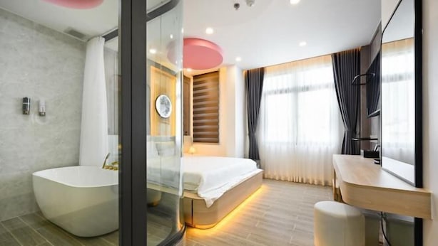 A modern bathroom with a freestanding bathtub, a glass shower enclosure, a bed with a wooden headboard, and a vanity with a mirror. - Lotus Airport Hotel Saigon (Ho Chi Minh City)